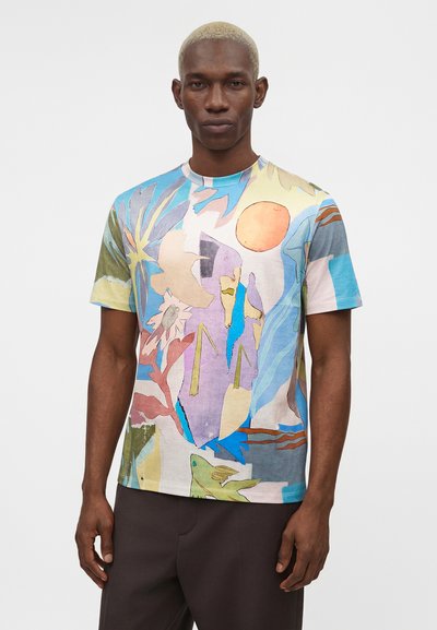 Άντρας με κοντά ξανθά μαλλιά φοράει πολύχρωμο μπλουζάκι με αφηρημένο σχέδιο που απεικονίζει σχήματα, φυτά, έναν ήλιο και ένα ψάρι σε μονόχρωμο φόντο.