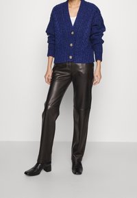 Marinblå stickad cardigan med grov textur, v-ringning, tre stora bruna knappar, bärs över svarta läderbyxor och ankelskor.