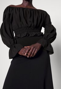 Blouse noire à épaules dénudées avec de longues manches bouffantes, taille froncée et accents volants. Tissu lisse au fini mat.