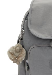 Sac à dos gris avec une poche frontale zippée et un porte-clés en forme de singe en peluche beige et doux attaché à la sangle latérale.