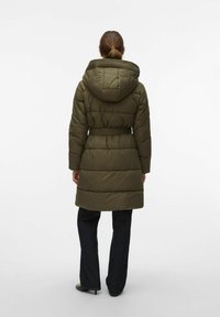 Ženska nosi olivnozeleno podolgo puhovko s kapuco in pasom, v kombinaciji s temnimi ravnimi hlačami in črnimi petkami, stoje obrnjena stran.