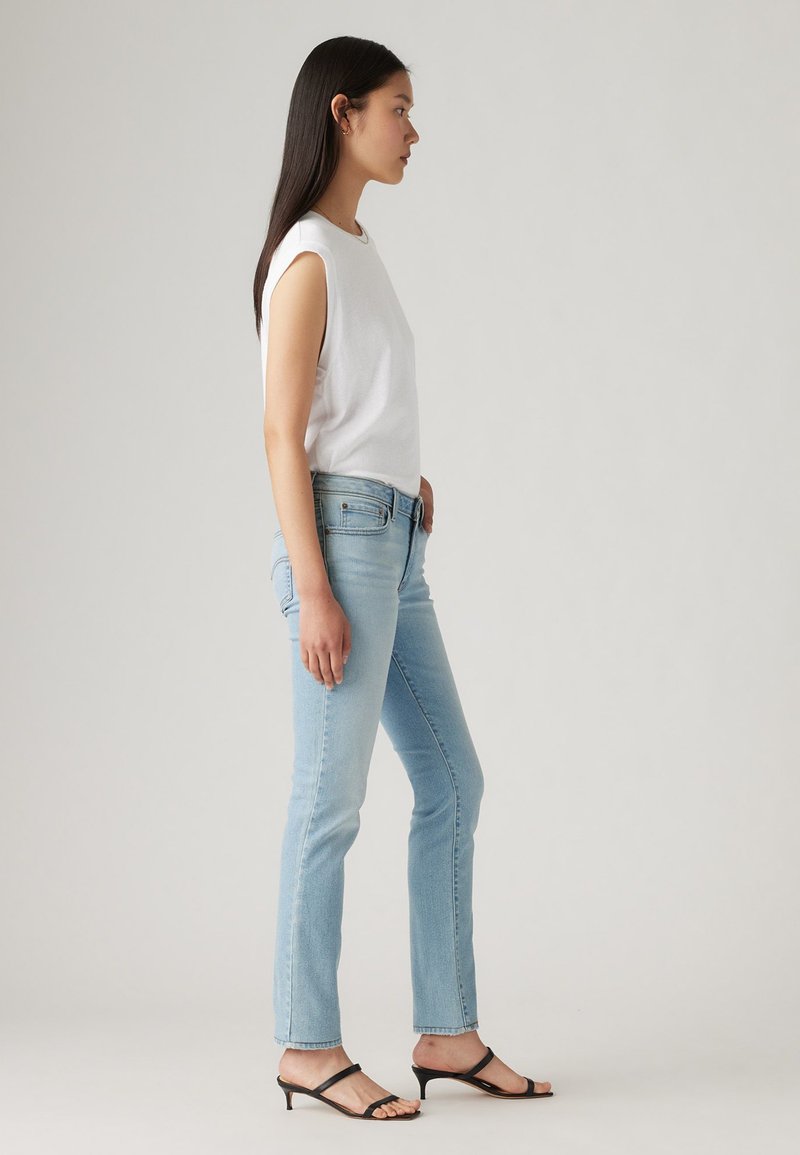 Top sans manches blanc, jeans bleu clair et sandales à talons noires. Jeans ajustés avec un délavage léger, texture lisse et détails minimaux.