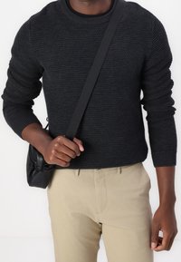 Ciemny sweter z grubym splotem o fakturowanej powierzchni, z okrągłym dekoltem i długimi rękawami, zestawiony z jasnobeżowymi spodniami i małą czarną torebką na ramię.