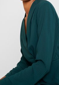 Grönblå omslagstopp med texturerad väv, med V-ringning och långa ärmar. Materialet har ett subtilt rutmönster.