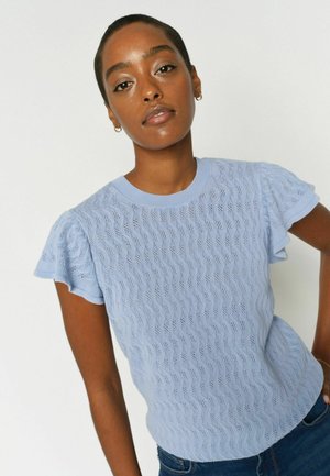 Femeie cu păr scurt, purtând un tricou texturat bleu cu mâneci scurte și blugi bleumarin, stând în fața unui fundal alb simplu.