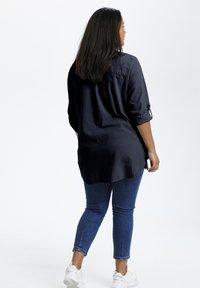 Chemise à manches longues navy en tissu léger, avec un ourlet arrondi et des manches retroussées. Couplée à un jean ajusté et des baskets blanches.