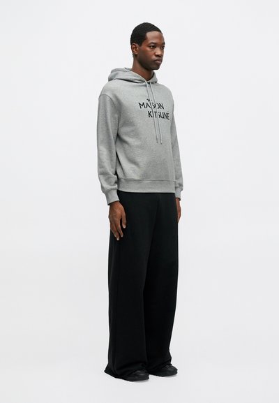 Hanorac gri cu glugă, prezentând un logo imprimat negru. Asortat cu pantaloni negri cu croială largă. Design simplu, aspect casual.