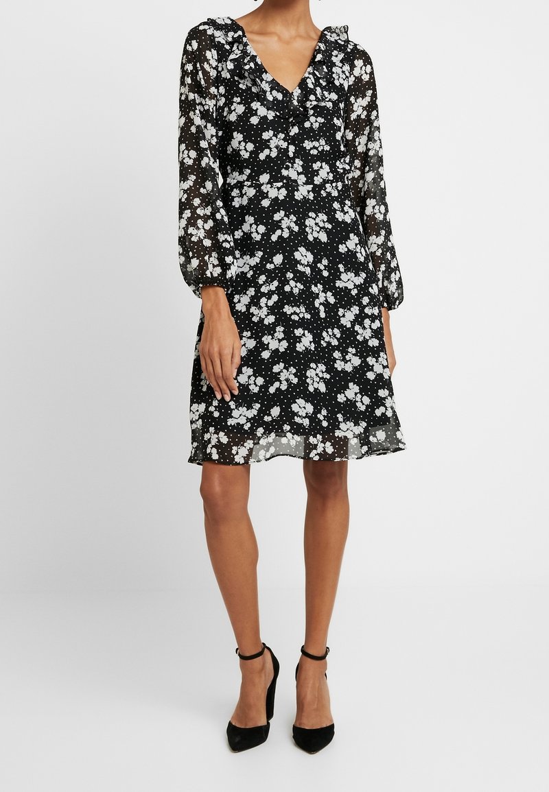 Femme portant une robe noire longueur genou avec imprimé floral blanc et manches longues transparentes, associée à des talons noirs à bride cheville.