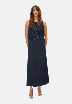 Donna che indossa un abito lungo blu navy senza maniche con inserti in pizzo floreale sul petto, abbinato a tacchi bianchi aperti sulle dita.