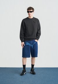 Νέος άνδρας που φοράει μαύρο φούτερ, μπλε τζιν σορτς με μεγάλο λογότυπο, μαύρα αθλητικά παπούτσια, μαύρες κάλτσες και σκούρα γυαλιά ηλίου, στέκεται σε μπλε χαλί.