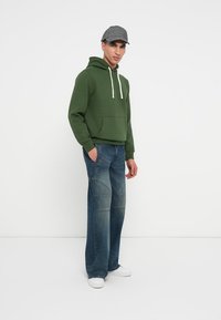 Zaļš kapuci krekls ar baltām auklām un priekšējo kabatu, apvienots ar gaiši ziliem džinsiem ar paneļu detaļām un baltām sporta kurpēm. Pelēks kapuce.