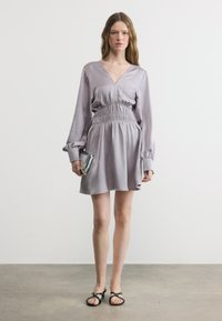 Γυναίκα όρθια, φοράει ένα ανοιχτό μωβ φόρεμα με μακριά μανίκια και σούρες στη μέση, κρατάει ασημένιο clutch και φοράει μαύρα σανδάλια με ανοιχτά δάχτυλα.