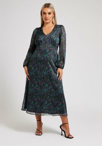 Lång svart klänning med veckad design som har lila och gröna blommönster, v-ringning och puffärmar, ihop med högklackade skor.