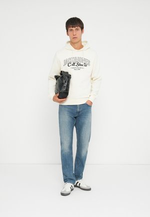 Sudadera blanca con texto negro, jeans azules y bolso negro. Zapatillas blancas con rayas negras y grises. Corte recto. Fondo liso.