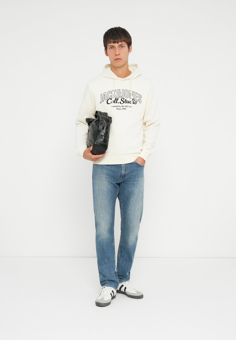 Hvid hoodie med sort tekst, blå jeans og sort taske. Hvide sneakers med sorte og grå striber. Lige pasform. Ensfarvet baggrund.