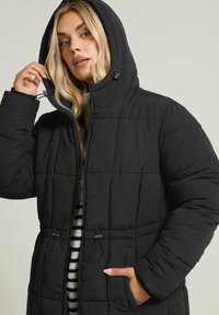 Czarna kurtka puchowa z kapturem, pikowana, z regulowanym paskiem w talii, kieszeniami z przodu i zamkiem błyskawicznym. Wyłożona w paskowaną podszewkę.