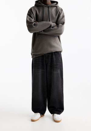 Harmaa huppari, jossa on etutasku, yhdistettynä väljien mustien farkkujen kanssa, joissa on hienovaraisia raidoituksia, ja valkoiset tennarit kumipohjilla.