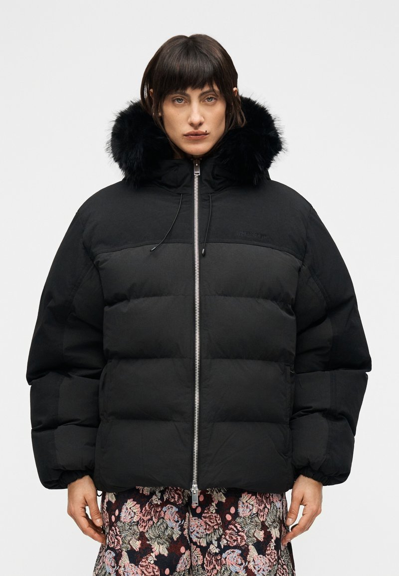 Μαύρο ανδρικό μπουφάν puffer με αποσπώμενη κουκούλα από συνθετική γούνα, με φερμουάρ μπροστά, ελεύθερη γραμμή στους ώμους και ριμπ μανσέτες. Υφή με καναλώσεις.