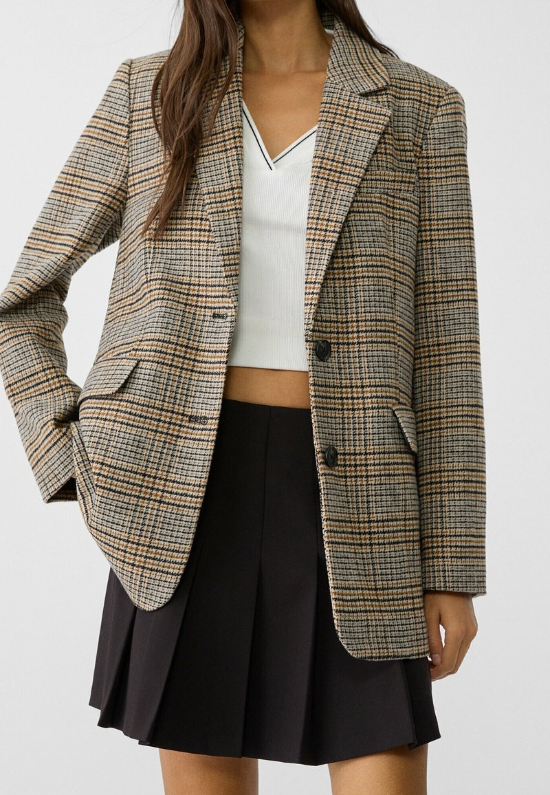 Femme portant un blazer à carreaux beige et marron, un crop top blanc à col en V et une jupe plissée noire, debout devant un fond uni.