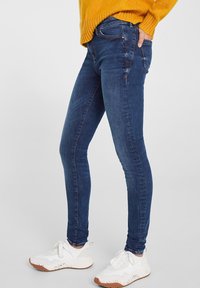 Skinny-fit denim jeans in dark blue, made of stretchy fabric. Features classic five-pocket design and subtle stitching details. Paired with white sneakers.