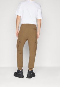 Bruna bomullscargopants med elastiska manschetter och flera sidofickor, bärs med en vit t-shirt och svarta sneakers, sedd bakifrån.