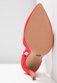 Chaussure à talon haut rouge à bout pointu vue de la semelle, avec un talon fin, appuyée contre un mur et une surface blancs.