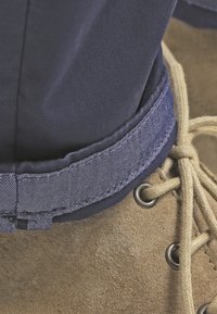 Jean en denim bleu retroussé au-dessus d’une botte en daim beige à lacets beige visibles à travers des œillets métalliques.