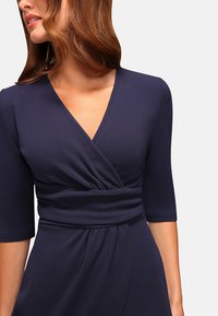 Femme portant une robe portefeuille bleu marine avec des manches trois-quarts et une taille froncée, vue du cou aux hanches devant un fond uni.