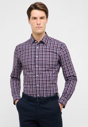 Homme portant une chemise à carreaux bleu marine, blanc et rouge, boutonnée et rentrée dans un pantalon bleu marine foncé, les mains dans les poches, sur un fond uni.