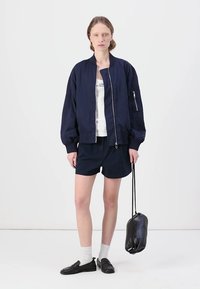 Ναυτική bomber ζακέτα με φερμουάρ μπροστά, ελαστικούς καρπούς και πλενόμενους γοφούς, συνδυασμένη με λευκό μπλουζάκι, ναυτικές σορτς και μαύρα παντόφλια.