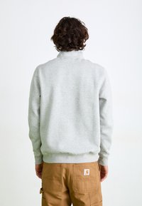 Grå fleece sweatshirt med høj krave, ribbede manchetter og talje. Synlig etiket på baglommen af brune arbejdsbukser.