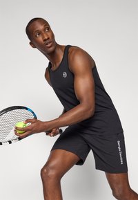 Svart athletic tank top, tettsittende, med en logo på brystet, kombinert med svarte shorts med hvit tekst. Holder en tennisketsj og en ball.