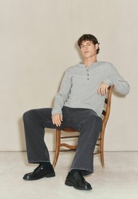 Grå langærmet henley-skjorte med knapper, sorte vide jeans og sorte polerede sko, poseret på en træstol mod en neutral baggrund.