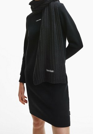 Mannequin portant une robe Calvin Klein noire à manches longues et une écharpe épaisse assortie en tricot, avec les logos Calvin Klein visibles sur les deux articles.
