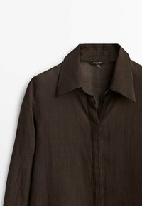 Brązowa lniana koszula z wywijanym kołnierzykiem, długimi rękawami i zapięciem na guziki z przodu. Tekstura wydaje się lekka i lekko pognieciona.
