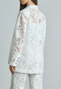 Completo bianco in pizzo con un intricato motivo floreale, composto da un blazer con colletto e bottoni e pantaloni coordinati, che mostrano un design testurizzato.