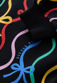 Tissu noir avec des motifs de rubans ondulés colorés et un détail de bord-côtes, avec un texte bleu clair partiellement visible indiquant "LITTLE B".