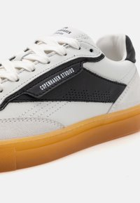 Valge nahast sneaker mustade aktsentidega, millel on tekstuuriga sametpaneel, lukkupandav disain ja kummine kummipõhi. Logo külgmisel osal.
