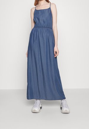 Vrouw die een lange blauwe mouwloze jurk met dunne bandjes en witte chunky sneakers draagt, staande tegen een effen lichte achtergrond.