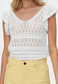 Biały koronkowy top z krótkimi falbaniastymi rękawami, charakteryzujący się otwartym wzorem dzianiny i falbaniastym dołem, w połączeniu z żółtymi spodniami o wysokim stanie.