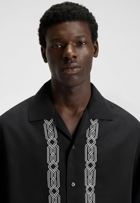 Camicia nera a bottoni con un'importante ricamo geometrico bianco sul davanti. Collo corto e vestibilità rilassata.
