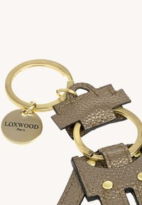 Trousse en or avec deux breloques en cuir marron texturé et une étiquette ronde en or gravée "LOXWOOD Paris" sur un fond clair.