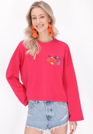 Rosa Sweatshirt mit langen Ärmeln, verziert mit floralen Stickereien. Kombiniert mit ausgefransten Denim-Shorts. Die Accessoires umfassen orangefarbene blütenförmige Ohrringe.