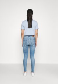 Camisa de color azul claro con manga corta combinada con jeans ajustados de mezclilla clara. El modelo lleva zapatillas deportivas blancas y tiene el pelo largo recogido en una coleta.