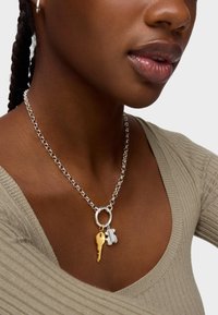 Collier en chaîne en argent comprenant un maillon circulaire et deux breloques : une clé en or et un cadenas en argent, porté contre un haut de couleur neutre.