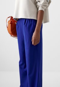 Labave, svijetloplave hlače s glatkom teksturom, u kombinaciji s laganim bež duksericom i narančastom torbom.