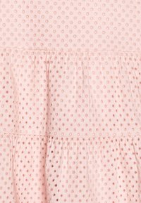 Lichtroze stof met een gestippeld patroon, voorzien van kleine uitgesneden cirkels. Het onderste gedeelte heeft geplooide ruches, die textuur en volume toevoegen.