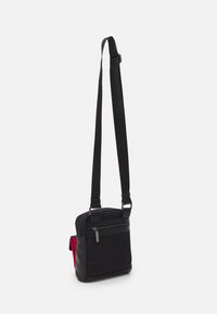 Borsa nera a spalla con tasca frontale con zip, materiale in nylon, forma rettangolare e accento laterale rosso. Tracolla regolabile per maggiore comodità.