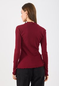 Dlhý rukáv top v burgundskej ribovanej látke, so riaseným výstrihom a lemom. Kombinovaný s čiernymi voľnými nohavicami.