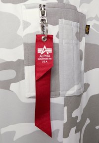 Röd tygnyckelring med "Alpha Industries Inc U.S.A." fäst vid en grå och vit camouflagejacka med dragkedjeficka.